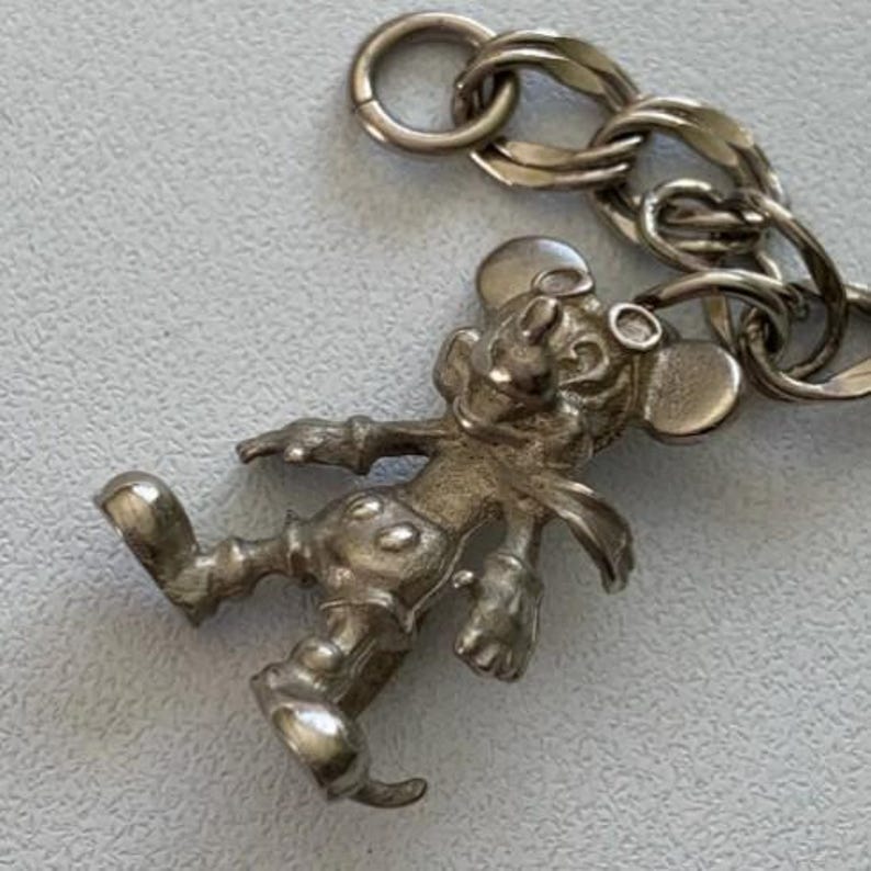 Puede incluir: Un dije plateado de Mickey Mouse con una cadena. El dije representa a Mickey Mouse en una pose caminando, con sus pantalones cortos y guantes caracter&iacute;sticos. La cadena est&aacute; hecha de eslabones entrelazados.