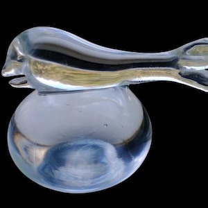 May include: A clear glass sculpture of a bird perched on a round base. The bird has a long, curved body and a detailed head with an open beak. The sculpture is set against a black background.