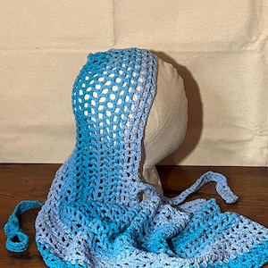 May include: A handmade crochet bonnet in shades of blue, transitioning from light to dark. The bonnet features an open, net-like weave and long ties for securing. The item is displayed on a mannequin head.