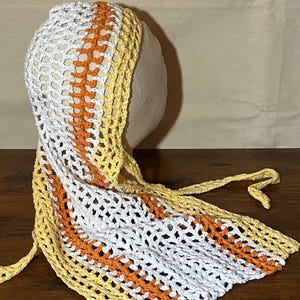 May include: A handmade crochet bonnet in white, orange, and yellow. The bonnet features a striped design with a tie closure. The open weave construction is visible throughout the bonnet. The bonnet is likely for a baby or child.
