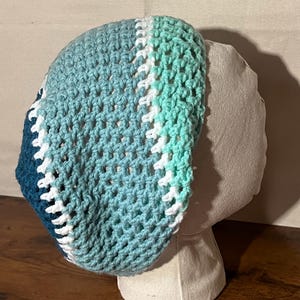 May include: A handmade crochet hat in shades of blue, teal, and white. The hat features a textured, open weave design. The hat is displayed on a mannequin head, showcasing its shape and style. The hat is a stylish accessory.