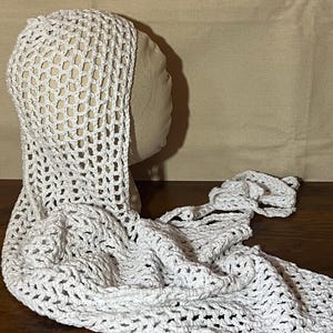 May include: A white, open-weave crochet hood and scarf set. The hood is draped over a mannequin head, with the scarf cascading down. The crochet pattern features a large, open stitch design, creating a delicate and airy appearance.