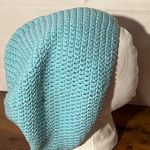May include: A light blue, handmade crochet beanie hat. The hat is displayed on a mannequin head. The hat is made of a soft, textured yarn and has a ribbed pattern. The beanie is a slouchy style.