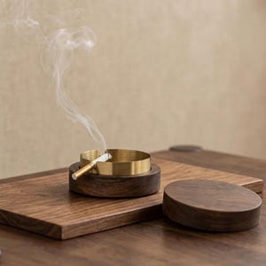 Puede incluir: Un cenicero de latón y madera con un cigarrillo encendido, emitiendo humo. El cenicero está colocado sobre una bandeja de madera, con una tapa de madera separada. El diseño es minimalista y moderno.