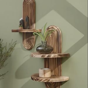 May include: Two wooden wall shelves with an arched design. The shelves are made of light brown wood and have a natural finish. One shelf holds a small plant in a glass vase, and a candle. The other shelf holds decorative stones.
