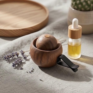 Puede incluir: Un ambientador de coche de madera con un difusor redondo en forma de bola y un clip negro. Una pequeña botella de vidrio con un cuentagotas y una tapa de madera está al lado. El ambientador está sobre una superficie gris.
