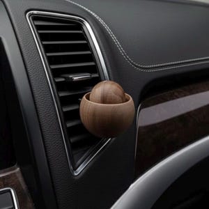 May include: A wooden car air freshener with a spherical design. The air freshener is made of light brown wood and is composed of a small sphere nested inside a larger, bowl-shaped piece. It is attached to a car's air vent.