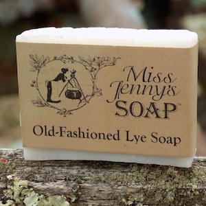 May include: A bar of white lye soap with a brown paper label. The label has a black and white illustration of a woman making soap and the text "Miss Jenny's SOAP" and "Old-Fashioned Lye Soap".
