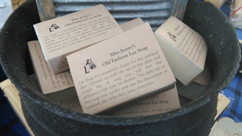 May include: A close-up of several brown cardboard boxes of Miss Jenny's Old Fashion Lye Soap. The boxes are stacked in a black cauldron. The soap is described as a wonderful choice for dry, irritated skin. The text on the boxes reads: "Lye Soap is a wonderful choice for dry, irritated skin. Chances are with the use of this plain and simple bar of soap you will feel the difference with your 1st shower. The daily use of this soap may soothe and ease the discomfort of irritated, dry, itchy skin, acne, or eczema."