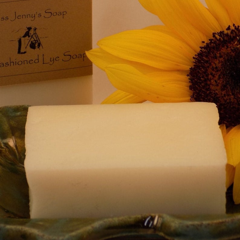 May include: A bar of white soap on a green and brown ceramic dish. A sunflower is in the background. The soap is labeled "Old Fashioned Lye Soap".