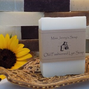 May include: A bar of white lye soap with a brown paper label that reads "Miss Jenny's Soap Old Fashioned Lye Soap". The soap is sitting on a brown and white patterned ceramic dish. A sunflower is in the background.