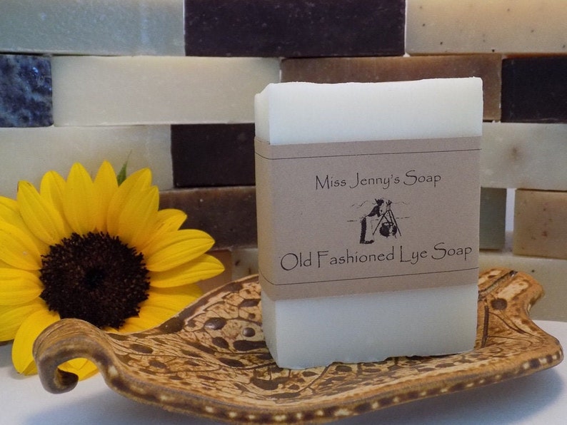 May include: A bar of white lye soap with a brown paper label that reads "Miss Jenny's Soap Old Fashioned Lye Soap". The soap is sitting on a brown and white patterned ceramic dish. A sunflower is in the background.