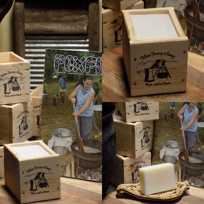 May include: A wooden box with a white label that reads "Miss Jenny's Soap" and a black and white illustration of a cauldron with a dog standing next to it. The text "Wash and be Cleaned" is printed below the illustration. The box is sitting on a wooden surface. There are other boxes in the background.
