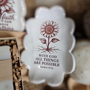 May include: White ceramic dish with a scalloped edge, featuring a brown sunflower illustration and the text "WITH GOD ALL THINGS ARE POSSIBLE" from Matthew 19:26. The dish has a decorative, floral shape.