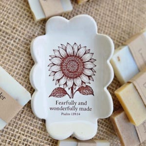 May include: White ceramic soap dish with a scalloped edge, featuring a detailed brown sunflower illustration and the text "Fearfully and wonderfully made Psalm 139:14." Several bars of soap are visible in the background.