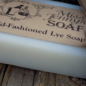 May include: A bar of white lye soap with a brown paper label that reads "Miss Jenny's Soap Old-Fashioned Lye Soap". The label has a vintage illustration of a woman making soap.