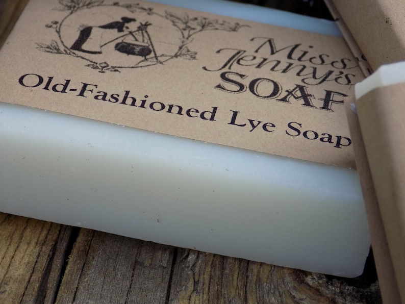 May include: A bar of white lye soap with a brown paper label that reads "Miss Jenny's Soap Old-Fashioned Lye Soap". The label has a vintage illustration of a woman making soap.