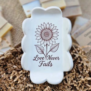 May include: White ceramic soap dish with a sunflower illustration and the words "Love Never Fails" in a decorative font. The dish has a scalloped edge and is surrounded by brown packing material.