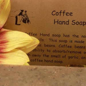 May include: A bar of brown coffee hand soap with a sunflower in the background. The text on the label reads "Coffee Hand Soap" and explains that the soap is made with coffee beans, which have the ability to absorb and remove odors.
