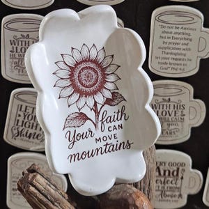 May include: White, scalloped-edge dish with a sunflower design in shades of brown. The dish features the text "Your faith can move mountains" in a decorative font. Surrounding the dish are other decorative items.