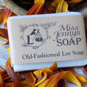 May include: A bar of white soap with a brown paper label that reads "Miss Jenny's SOAP" and "Old-Fashioned Lye Soap". The label also features a black and white illustration of a woman making soap.