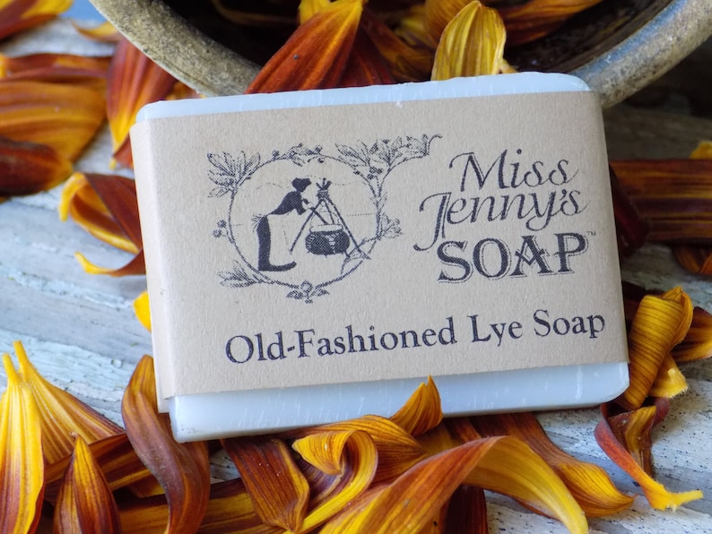 May include: A bar of white soap with a brown paper label that reads "Miss Jenny's SOAP" and "Old-Fashioned Lye Soap". The label also features a black and white illustration of a woman making soap.