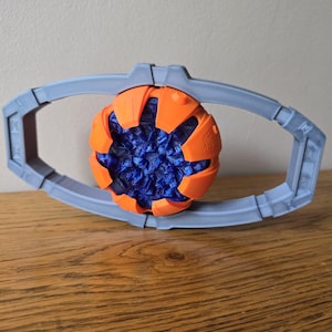 以下が含まれることがあります： オレンジ色の外観と青色の結晶状の内部を持つ、3Dプリントされた未来的な外観のオーブ。オーブは、薄いグレーの楕円形のフレーム内に保持されています。木製の表面に表示されています。