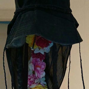 Può includere: Cappello nero con tesa larga e velo nero trasparente. Una ghirlanda di fiori artificiali colorati, tra cui rose rosa, gialle e rosse, pende dal cappello. Il cappello ha dei cordini neri.