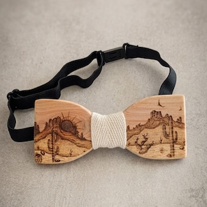 Peut inclure: Nœud papillon en bois avec un motif de paysage désertique. Le bois clair présente une scène gravée au laser de cactus, de montagnes et d'un soleil. Une bande tissée crème entoure le centre et une sangle noire réglable complète le design.