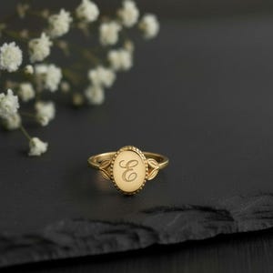 Puede incluir: Un anillo de sello dorado con una cara ovalada, con la letra "E" grabada. El anillo tiene un borde con cuentas y detalles de hojas en los lados. El anillo se muestra sobre una superficie oscura.
