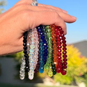May include: A hand holds a collection of beaded bracelets in a rainbow of colors. The bracelets feature faceted beads in shades of black, clear, pink, blue, green, yellow, orange, and red. The bracelets are strung together, creating a vibrant display.