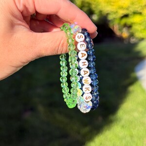 May include: Three beaded bracelets in shades of green and blue are held up. One bracelet spells out "SEAHAWKS" with white letter beads. The bracelets are made of clear, faceted beads. The background is blurred green grass.