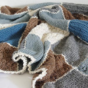 Puede incluir: Una manta de punto con diseño de patchwork. La manta presenta cuadrados azules, marrones, grises y blancos. Los bordes están acabados con un ribete blanco festoneado. La manta está doblada y drapeada, mostrando la textura y las variaciones de color.