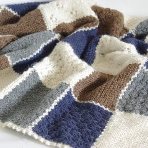 Könnte beinhalten: Eine gestrickte Decke mit einem Patchwork-Design aus quadratischen Blöcken in Marineblau, Creme, Braun und Grau. Die Quadrate wechseln zwischen glattem Rechtsstrick und strukturierten Mustern, verbunden durch eine weiße gehäkelte Umrandung.