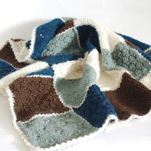Könnte beinhalten: Eine gehäkelte Decke mit einem Patchwork-Design aus quadratischen Blöcken in den Farben Dunkelblau, Hellblau, Braun und Creme. Die Decke ist mit einem strukturierten Stichmuster gefertigt und mit einem cremefarbenen Häkelrand versehen.