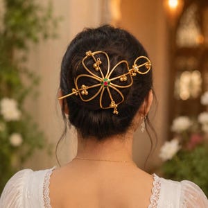Op de afbeelding: Een goudkleurig haaraccessoire in de vorm van een bloem, versierd met kleine gouden kralen en een centrale edelsteen met rode en groene accenten. Het haarstuk wordt in een knot gedragen.
