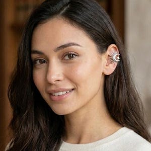 Puede incluir: Un pendiente de oreja plateado con un diseño de sol con una luna creciente y una estrella. La joya se lleva en la oreja de una persona con cabello oscuro y un suéter blanco. El pendiente añade un acento celestial a la oreja.