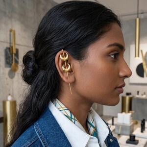 May include: Gold-toned ear cuff and earring set. The ear cuff features two crescent moon shapes, and the earring has a long, straight drop. The jewelry is worn on the ear of a person with dark hair and a denim jacket.