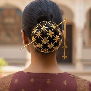 Könnte beinhalten: Eine Frau trägt einen dekorativen goldenen Haarschmuck. Das Accessoire hat ein kugelförmiges Käfigdesign mit floralen Ausschnitten, einen langen Stift und baumelnde Kettenakzente. Die Frau trägt ein bordeauxfarbenes Oberteil mit goldenen Blumenmustern.