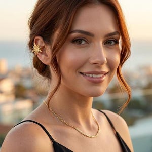 Può includere: Una donna indossa un orecchino a forma di stella dorata e una delicata collana a catena dorata. Indossa un top nero con spalline sottili. Lo sfondo è sfocato, con vista sull'oceano e sugli edifici.