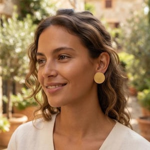 May include: A gold-toned circular stud earring with a textured, matte finish. The earring is round and flat, featuring a simple geometric design that sits flush against the earlobe. The surface has a subtle, granular appearance.