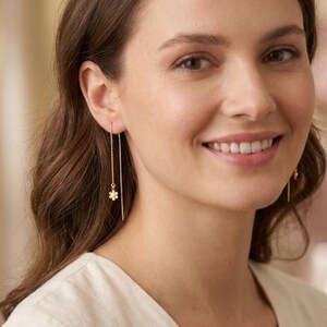 May include: A pair of delicate gold earrings. Each earring features a fine chain that dangles from the ear, with a small, white flower charm at the end of one chain. The earrings are a simple and elegant design.