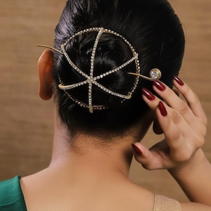 Könnte beinhalten: Ein goldfarbenes Haaraccessoire mit einem kreisförmigen Design, verziert mit kleinen, funkelnden Kristallen. Das Haarteil wird mit einer langen, schlanken Nadel befestigt. Das Haar der Person ist dunkel, und die Nägel sind tiefrot lackiert.