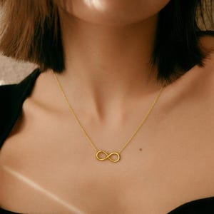 May include: A gold-coloured necklace with an infinity symbol pendant. The pendant is suspended from a fine chain, creating a minimalist design. The necklace is worn around the neck, against a dark background.