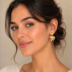 Peut inclure: Boucles d'oreilles dorées en forme de cœur avec des pierres précieuses colorées. Les boucles d'oreilles ont une petite boule dorée en haut et sont ornées de pierres rouges, bleues, vertes et jaunes. Portées par une personne aux cheveux foncés.