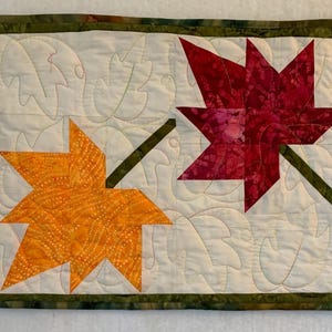 May include: A quilted table runner with a white background and four colorful maple leaves. The leaves are green, orange, red, and brown.