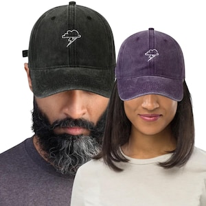 Puede incluir: Dos gorras de béisbol, una negra y otra morada, cada una con un diseño bordado en blanco de nube y rayo. Las gorras tienen un aspecto vintage y desgastado y correas ajustables.