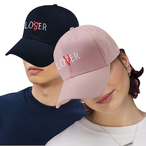 May include: Two baseball caps, one navy blue and one light pink. Both caps have the word "LOSER" embroidered in white, with the "O" replaced by a red heart. The caps have a curved brim and an adjustable strap.