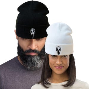 May include: Two knit beanies, one black and one white, each featuring a Scream mask logo embroidered on the front. The beanies are shown on two people. The black beanie is on a person with a beard. The white beanie is on a person with dark hair.