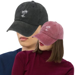 May include: Two baseball caps, one charcoal gray and one maroon, are displayed. Each cap features a white embroidered palm tree design on the front. The caps have a classic, slightly worn look, with a curved brim and adjustable strap.